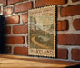 A vintage-style postage stamp from Maryland, depicting a scenic countryside landscape with a bridge and a house, is displayed on a brick wall.