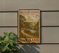 A decorative metal sign with a scenic countryside image of a bridge and house, mounted on a wall next to a plant.