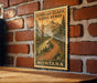 A framed illustration of a scenic mountain landscape with a winding road, mounted on a brick wall.