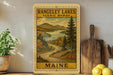 a framed vintage-style illustration of a scenic landscape with a winding road, mountains, and pine trees, along with the text "RANGELEY LAKES SCENIC BYWAY MAINE".