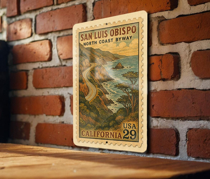 A vintage postage stamp featuring a scenic coastal landscape with a winding road, hanging on a brick wall.