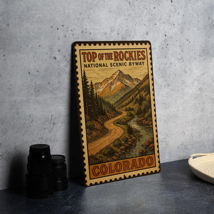 A vintage-style metal sign with a mountain landscape and a winding road, advertising "Top of the Rockies National Scenic Byway" in Colorado.