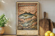 a framed stamp-like artwork depicting a fish, with a wooden cutting board and a potted plant in the background.