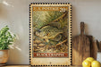 A framed U.S. postage stamp depicting a black crappie fish, placed on a wooden surface next to a wooden cutting board and a potted plant.