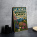 A vintage-style metal sign for the Alaska Botanical Garden, featuring illustrations of flowers and plants, is displayed on a gray concrete surface.