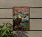A metal sign with a bald eagle perched on a tree branch, set against a backdrop of mountains and a river, with the text "ALASKA CHILKAT" and "BALD EAGLE PRESERVE" prominently displayed.