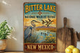 A vintage-style metal sign for Bitter Lake National Wildlife Refuge in New Mexico, featuring an illustration of a heron and ducks in a lake surrounded by mountains.