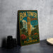 A vintage-style metal sign for the Bok Tower Gardens in Florida, featuring a tropical landscape with palm trees and a clock tower.