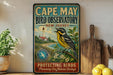 A vintage-style metal sign for Cape May Bird Observatory in New Jersey, featuring a colorful illustration of a bird perched on a branch with a lighthouse in the background.