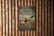 A metal sign with a vintage design hangs on a wooden wall, featuring an image of a deer and an eagle.