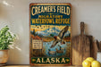 A vintage-style metal sign advertising Creamer's Field, a migratory waterfowl refuge in Alaska, with a scenic illustration of ducks flying over a lake.