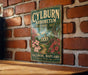 A vintage-style metal sign for the Baltimore Arboretum, featuring a scenic illustration of a garden with a house and flowers.
