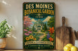 A vintage-style metal sign for the Des Moines Botanical Garden in Iowa, featuring a colorful illustration of a greenhouse surrounded by flowers and plants.