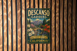 A vintage-style metal sign for Descanos Gardens, featuring a scenic landscape with a bridge and flowers, is displayed on a wooden wall.