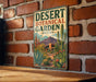 A vintage-style metal sign for the Desert Botanical Garden in Arizona, featuring a desert landscape with cacti, succulents, and a mountain backdrop.