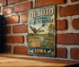 A vintage-style metal sign for the Desoto National Wildlife Refuge in Iowa, featuring an illustration of a goose in flight over a grassy field.