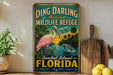 A vintage-style metal sign for Ding Darling National Wildlife Refuge in Sanibel Island, Florida, featuring a pink flamingo and a dolphin in a tropical landscape.