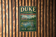 A vintage metal sign for Duke Farms, featuring a scenic landscape with a lake, trees, and a bird, is displayed on a wooden wall.
