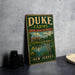 A vintage-style sign for Duke Farms, featuring a scenic landscape with a castle, river, and birds, is displayed on a gray countertop next to a stack of black camera lenses.