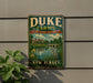 A vintage-style metal sign for Duke Farms, featuring a scenic landscape with a lake, bridge, and birds, mounted on a gray wall.