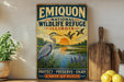 a vintage-style sign for the Emiquon National Wildlife Refuge in Illinois, with a scenic illustration of a heron and ducks in a lake. The sign also includes the text "PROTECT - PRESERVE - ENJOY A HAVEN FOR WILDLIFE" and is displayed on a shelf next to a wooden cutting board and some potted plants.