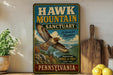 A vintage-style metal sign for the Hawk Mountain Sanctuary in Pennsylvania, featuring an illustration of a hawk in flight and text promoting the sanctuary as a haven for raptors.