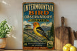a vintage-style metal sign for the Intermountain Bird Observatory in Idaho, with a colorful illustration of a bird and a mountain landscape.