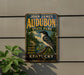 A vintage-style metal sign for John James Audubon State Park in Kentucky, featuring a black and white bird and text promoting the park's nature, history, and beauty.