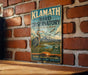 A vintage-style sign for Klamath Bird Observatory, featuring an illustration of a heron and mountains in the background, mounted on a brick wall.