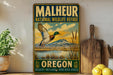 A vintage-style metal sign for the Malheur National Wildlife Refuge in Oregon, featuring an illustration of a duck in flight over a scenic landscape.