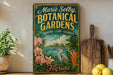 A vintage-style metal sign for "Marie Selby Botanical Gardens" in Florida, featuring a tropical landscape with palm trees, a bird, and flowers.