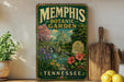 A vintage-style metal sign for the Memphis Botanic Garden in Tennessee, featuring a lush garden scene with a greenhouse and colorful flowers.