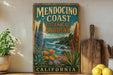 a vintage-style metal sign for Mendocino Coast Botanical Gardens in California, with a coastal landscape and colorful flowers on the sign.