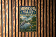 A vintage-style metal sign for the Minnesota Valley National Wildlife Refuge, featuring a heron and a scenic landscape, is displayed on a wooden wall.