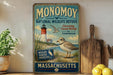 a vintage-style metal sign with a coastal scene, including a lighthouse, a bird, and a beach. The sign is titled "MONOMOY NATIONAL WILDLIFE REFUGE" and "CONSERVING COASTAL WILDLIFE TOMORROW". The sign also includes a message about wildlife habitat protection in Massachusetts.