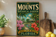 A vintage-style metal sign for the Mounts Botanical Garden in West Palm Beach, Florida, featuring a tropical garden scene with palm trees, flowers, and a gazebo.
