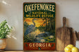a vintage-style sign for the Okefenokee National Wildlife Refuge in Georgia, with a painting of a heron and a sunset landscape.