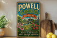 A vintage-style metal sign for "Powell Gardens" in Missouri, featuring a garden scene with flowers and a waterfall. The sign is displayed on a shelf next to a wooden cutting board and some potted plants.