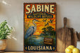 A vintage-style metal sign for Sabine National Wildlife Refuge in Louisiana, featuring an illustration of a blue heron and birds in flight against a sunset landscape.