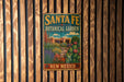 A vintage-style metal sign for the Santa Fe Botanical Garden in New Mexico, featuring a colorful illustration of a desert landscape with cacti, flowers, and a building.