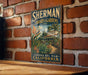 A vintage-style metal sign for Sherman Library & Gardens in Corona, California, featuring a tropical garden scene with palm trees and other plants.