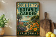 A vintage-style metal sign for the South Coast Botanic Garden in California, featuring a tropical garden scene with palm trees, flowers, and a path leading to the garden.