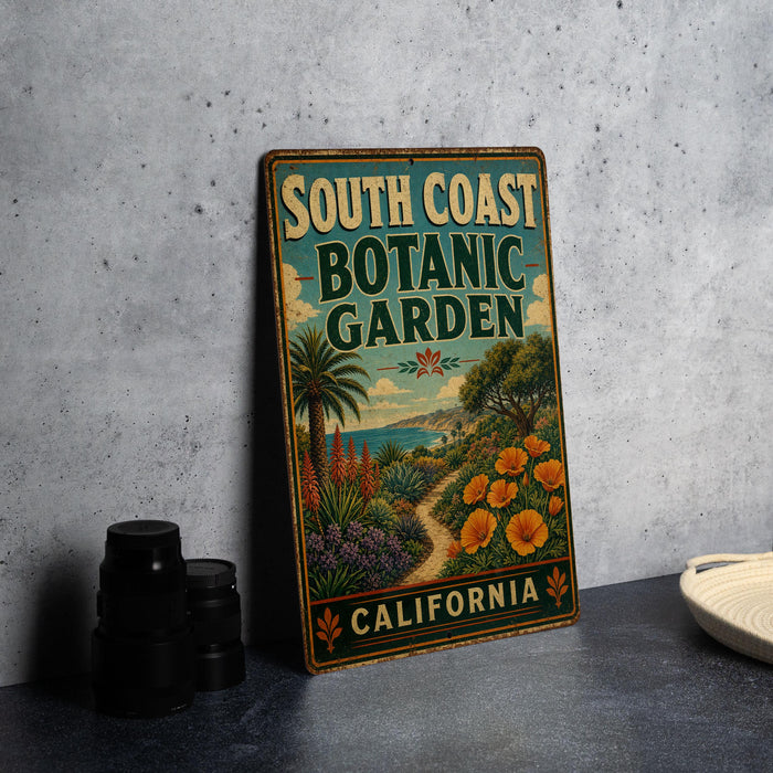 A vintage-style metal sign for the South Coast Botanic Garden in California, featuring a tropical garden scene with palm trees, flowers, and a winding path.