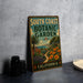 A vintage-style metal sign for the South Coast Botanic Garden in California, featuring a tropical garden scene with palm trees, flowers, and a winding path.