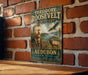 A vintage-style metal sign featuring a portrait of Theodore Roosevelt, the 26th President of the United States, and an illustration of a deer and an eagle, all set against a rustic brick wall.
