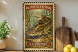 A vintage-style postage stamp featuring a flathead catfish, with the text "Flathead Catfish 15¢" and "U.S. Postage" printed on it. The stamp is displayed on a shelf next to a wooden cutting board and a potted plant.