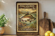 a framed print of a pink salmon, a type of fish, with a stamp-like border and a vintage-style illustration. The print is displayed on a wooden surface, with a potted plant, a cutting board, and some lemons nearby.