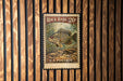 A vintage-style stamp featuring a bass fish, with the text "Rock Bass 20¢" and "U.S. POSTAGE" printed on it. The stamp is displayed on a wooden wall with vertical slats.