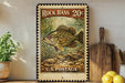 A vintage-style stamp featuring a large bass fish, with the text "Rock Bass 20¢" and "U.S. Postage" printed on it. The stamp is displayed on a shelf next to a wooden cutting board and a potted plant.