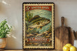 A vintage-style stamp depicting a spotted bass fish, with the text "5¢ SPOTTED BASS" and "U.S. POSTAGE WARM WATER CAME FISH" printed on it. The stamp is displayed on a shelf next to a wooden cutting board and a potted plant.